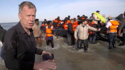 The BBC's Andrew Harding wearing a black shirt, standing in the sea, with a 'taxi-boat' full of migrants in the background. 