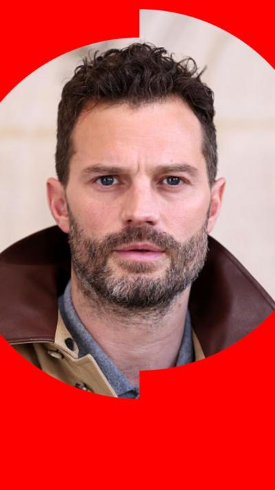 Jamie Dornan is wearing a beige jacket with a brown collar, and looks directly at the camera.