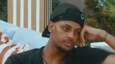 A man wearing a backwards cap rests his head on his hand while sitting on a couch.