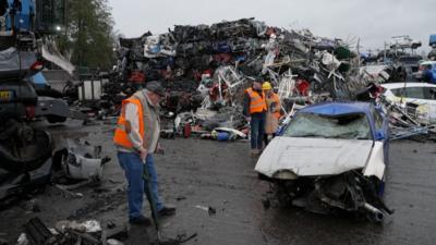 Three people in a scrapyard looking at a smashed-up car