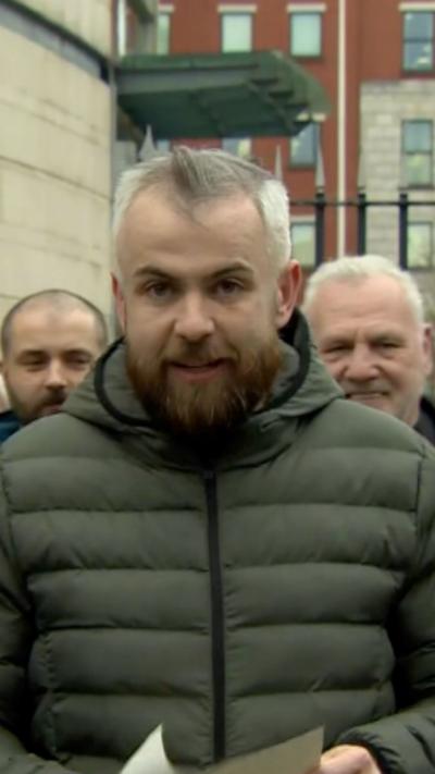Niall McNally reading a statement outside court. He is wearing a green puffer jacket. He has grey hair and a brown beard.