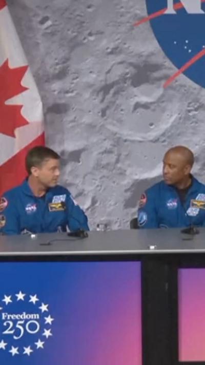 Two astronauts sit on a blue gradient table speaking into microphones with an image of the Moon and a Canadian flag in the background.