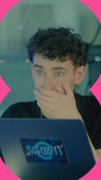 Man sat in front of a laptop holds his hand against his mouth. He looks shocked.