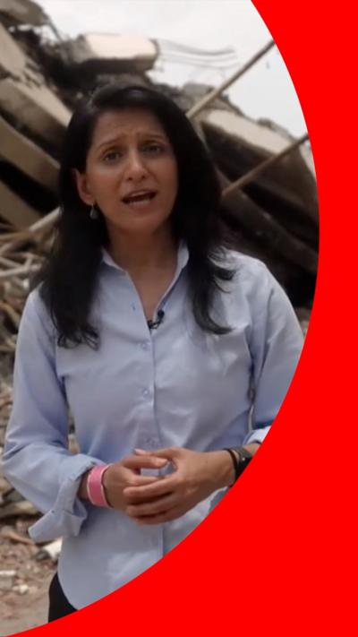 Yogita wears a blue shirt and speaks to the camera as she stands in front large piles of rubble, including what looks like a collapsed apartment block.