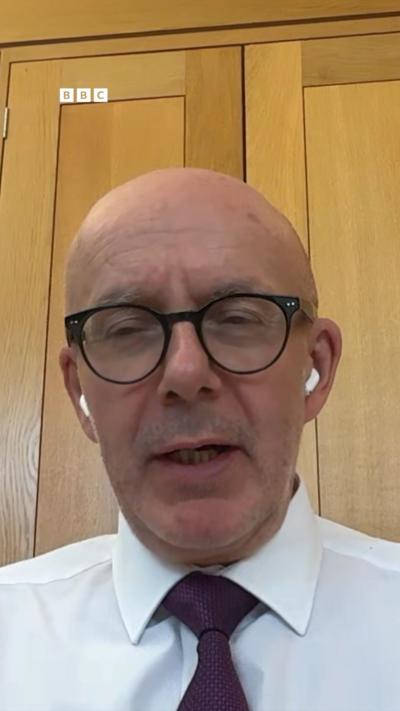 A bald man with black glasses and white shirt and claret tie sat in an oak panelled room