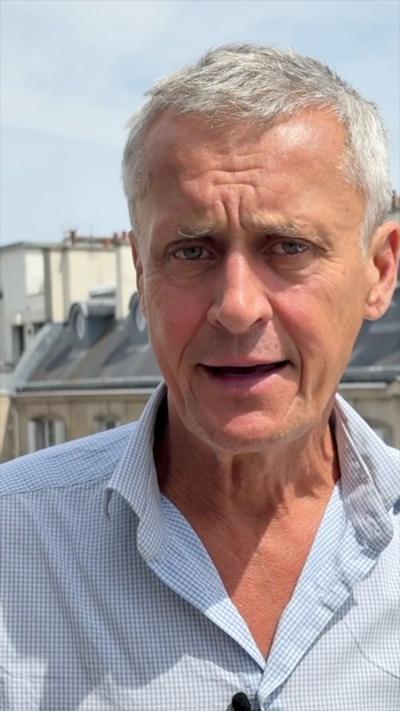 Hugh looks into the camera while standing in front of a row of houses