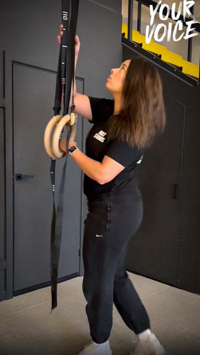 A woman with brown hair and wearing black t shirt and tracksuit bottoms moves gym equipment