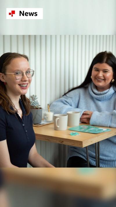 Two women sit either side of a table. They are both smiling at the camera. There are mugs between them and a plant between them.