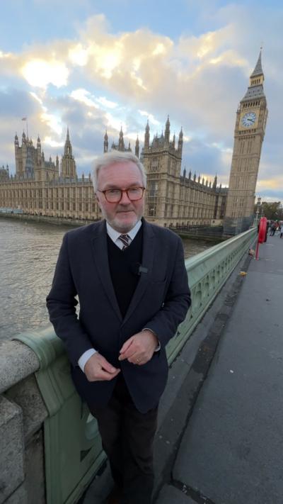 Political Editor Peter Henley wearing a dark suit and a shirt and tie standing on Westminster Bridge over the River Thames in London with the houses of Parliament and the Queen Elizabeth clock tower behind him. He is wearing square dark rimmed glasses.