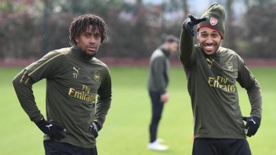 Alex Iwobi and Pierre-Emerick Aubameyang stand together on a green grass Arsenal training pitch, with Aubameyang, who is wearing an Arsenal woolly hat, pointing and pulling a silly face