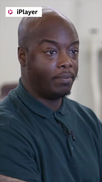 A man, Denzil Larbi, wears a dark green polo shirt and looks at someone off camera.