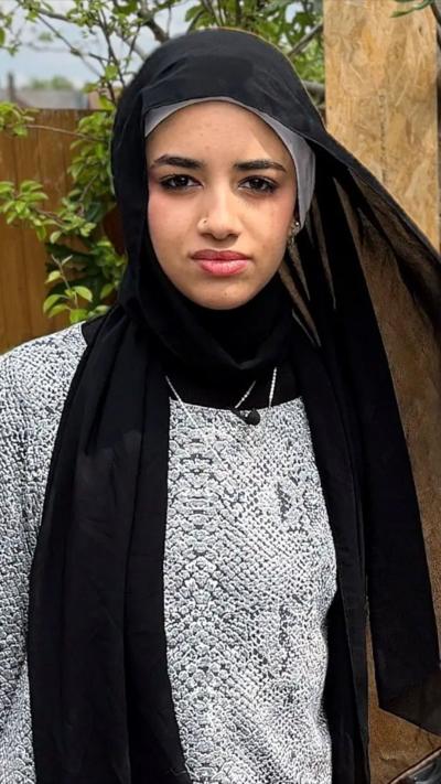 A lady wearing a black headscarf and grey jumper looks directly at the camera. Behind her is a wooden fence and branches and leaves of a tree