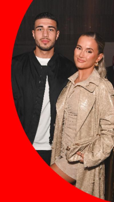 Tommy Fury and Molly-Mae-Hague stand next to each other and smile at the camera. Tommy wears a black jacket and white T-shirt while Molly-Mae wears a beige dress and jacket.
