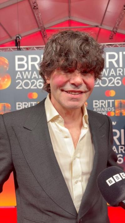 A man on the Brit Awards red carpet. He has curly brown hair that covers his eyes. 