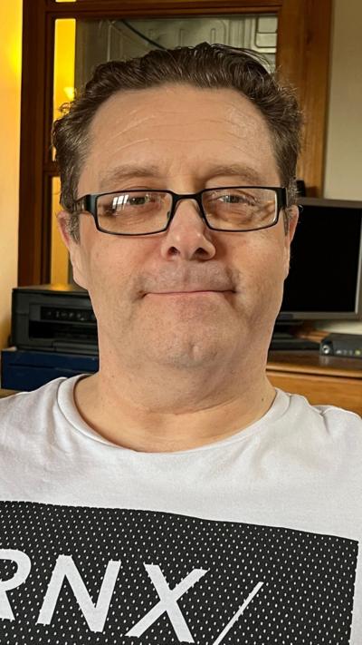 Mike Williams sitting in his living room in Caterham wearing a white tee shirt and spectacles.