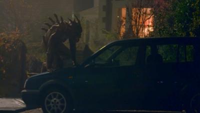 Shadowy monster with spikes sticking out of back crawls up a car in front of a house during the night.