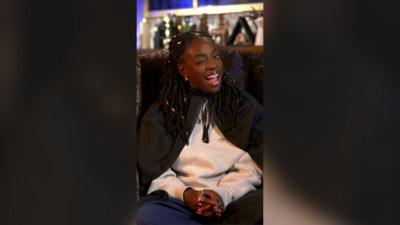 Cat Burns winks while sat in an armchair wearing a dark green cloak over a grey sweater. She has long black braids