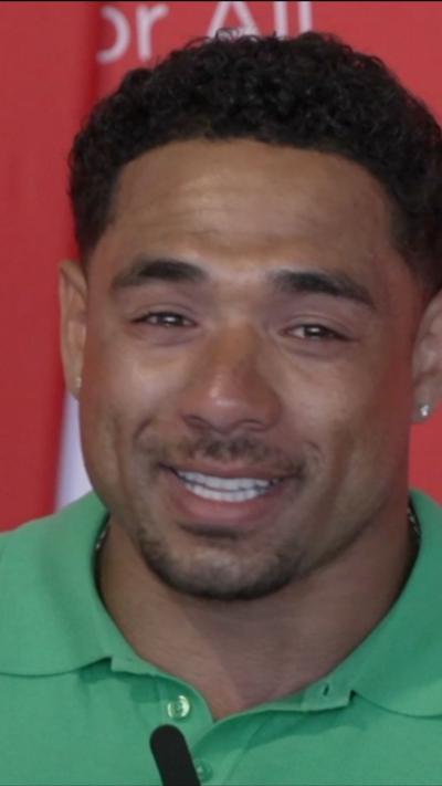A man in a light green polo shirt is smiling while looking at the camera. There are tears in his eyes. The background is a red wall with writing on it.