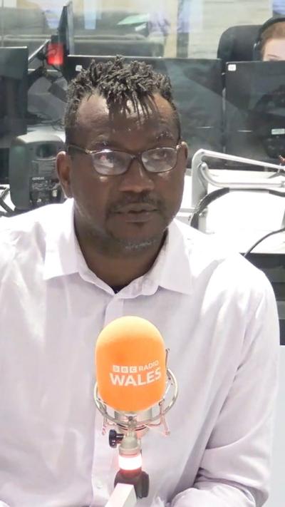 Joseff Gnagbo who wears glasses, sat in a radio studio behind an orange microphone, wearing a pink shirt