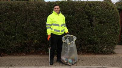 A man, Connor, stands on a pavement. He wears a hi-vis jacket and holds a litter picker and rubbish bag