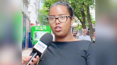 Woman stands on street and talks into BBC microphone that is being held in front of her