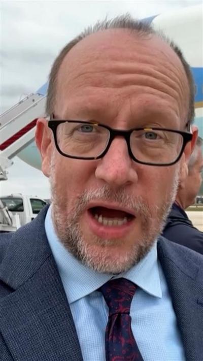 Tom Bateman, wearing a suit and black-rimmed glasses, speaks into the camera.