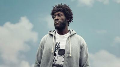 Stormzy as 'Tenzman' in Big Man. Wearing an afro wig and a white t-shirt and grey hoodie.