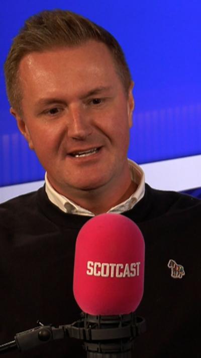 Man in a dark jumper and white shirt sitting behind a red radio microphone with Scotcast on it.