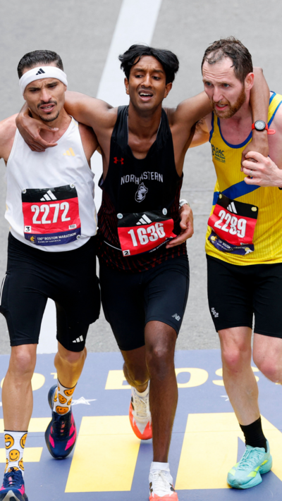 Three men wearing black shorts and different coloured running vests jog towards the finish line of the Boston Marathon with their arms around each other. Robson De Oliveira, who has short, dark hair and a moustasch and is wearing a white vest and a white headband. Ajay Haridasse has short, dark hair and a black vest. Aaron Beggs has short brown hair and a beard and is wearing a yellow vest with a blue stripe.