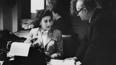 Black and white image of a woman working at a typewriter with a man leaning on her desk