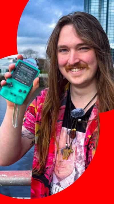 A man with long hair, wearing a brightly coloured pink shirt, holding up a green Gameboy