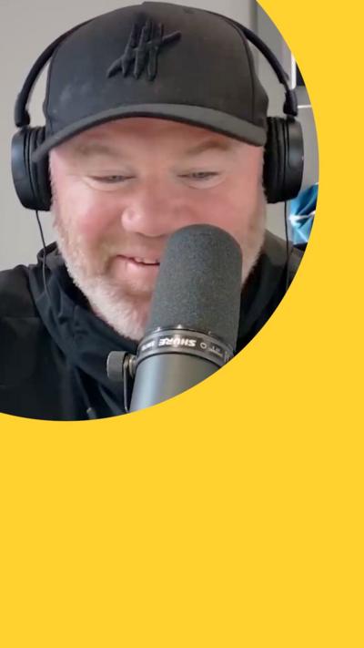 Wayne Rooney smiles behind a mic. He wears a black baseball cap and black headphones