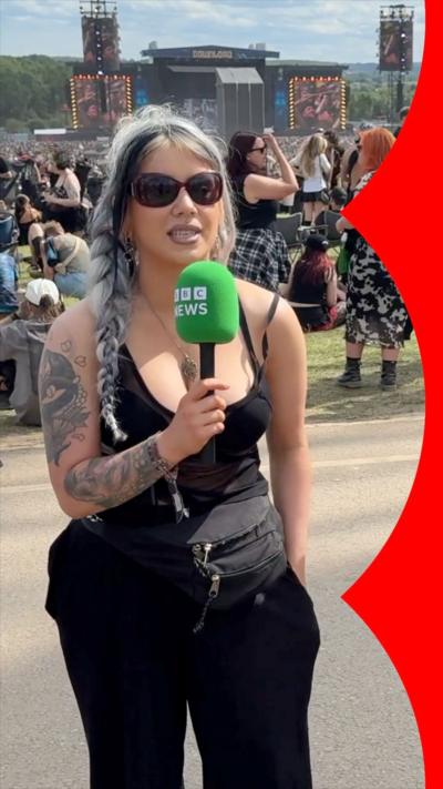 A woman stands in front of a festival stage and crowd. She has long silver and black hair, and wears a black sleeveless top, black jeans and sunglasses 