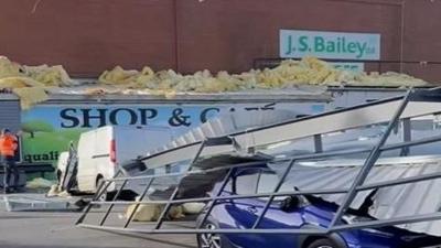 A blue car trapped underneath a large metal section of roof, with a brick-built building in the background. There is signage which read JS Bailey Ltd.