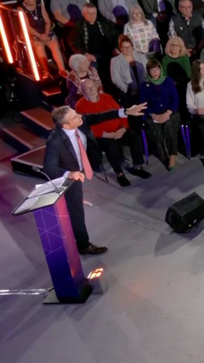 Nick Servini stood next to a lectern in front of a studio audience pointing