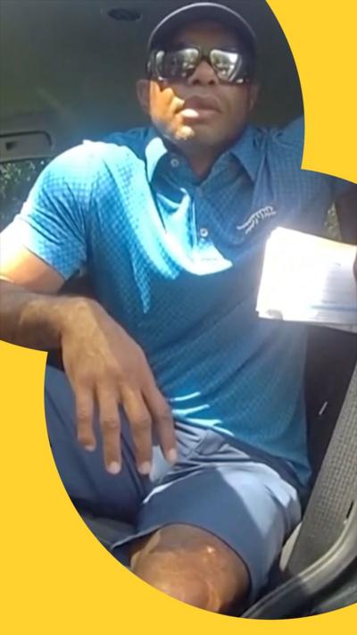 Tiger Woods sits in the driver's seat of his car. He is wearing a cap, sunglasses, slate-grey shorts and light blue polo shirt.