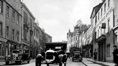 Black and white photograph of old cars driving down a street made of stone buildings.