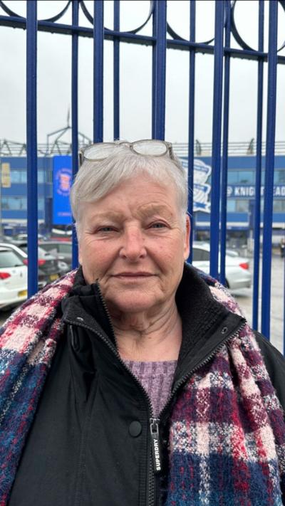 a woman with grey hair and glasses on her head stood in front of blue railings at a football stadium. She is wearing a black coat and a multi coloured scarf with blue, purple and pink sections