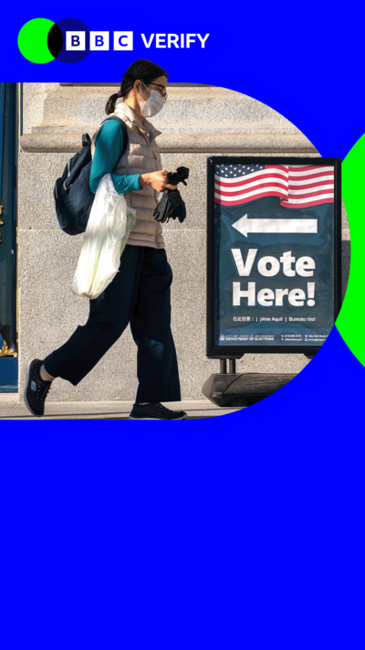 A woman wearing a face mask and carrying a shopping bag walks past a sign with an American flag on it saying "Vote Here!" 