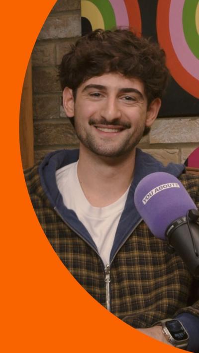 A man with short brown hair wears a checked jacket and sits in front of a purple microphone