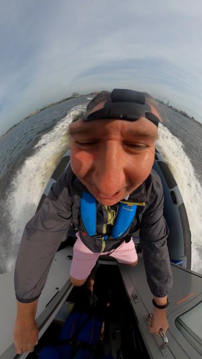 A distorted image of a man wearing a 360-degree camera from above on a boat with water around him