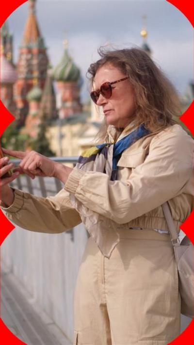 A woman in a beige mac and sunglasses uses her phone in front of the Kremlin.
