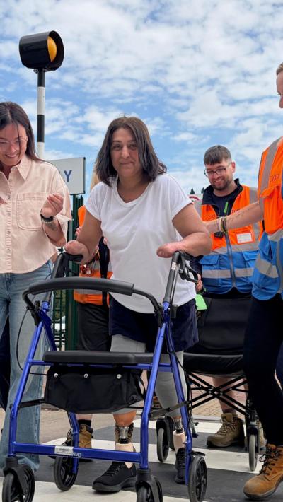 A woman in a white t-shirt with prosthetic limbs walks over a pelican crossing flanked by two women. One is wearing a pink shirt and jeans, and the other on the right is wearing black trousers and an orange and blue hi-vis jacket