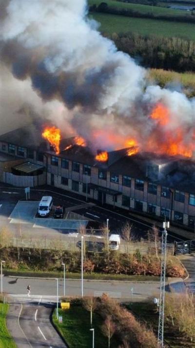 A building on fire. There are large clouds of smoke and orange flames coming out of the roof of the building. In front of the building is a car park and road. Behind the building is a field. 