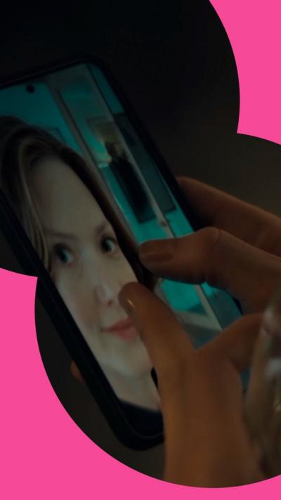 A hand pinches on a selfie on a phone screen to zoom in. A figure stands behind.