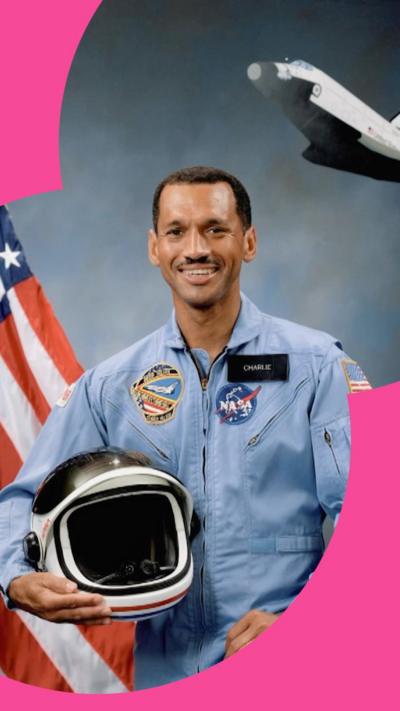 Charles Bolden poses in a blue Nasa uniform, holding a helmet. He stands in front of a US flag and an image of a space shuttle