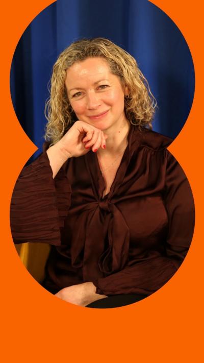 A woman with blonde curly hair and a maroon top, rests her head on her hand and smiles.