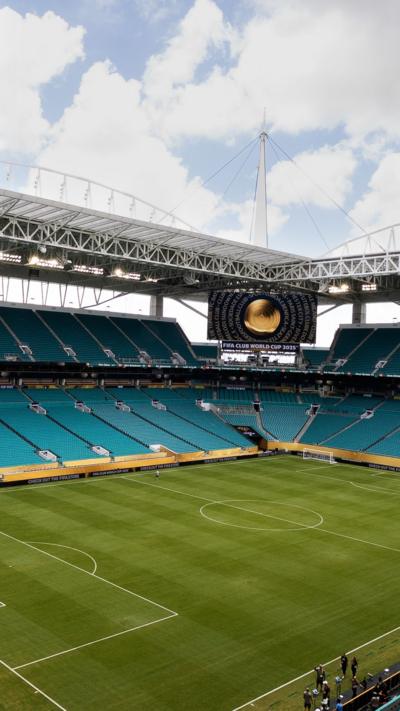 Empty football stadium, with green pitch and rows of turquoise seats and roofing.