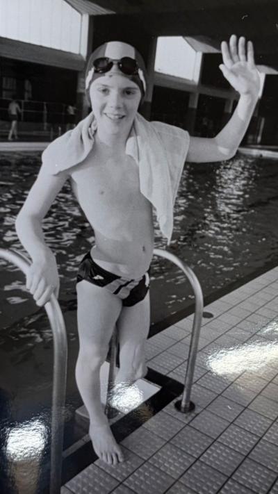A black and white image of a boy waving, with a towel on his left shoulder, standing on the edge of a swimming pool.