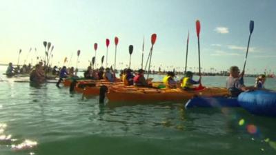 People in various types of small water craft holding their paddles upright in a Hervey Bay ceremony to remember whales killed by hunters